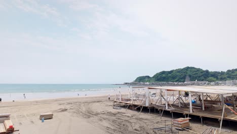 static wide shot of wooden beach structure under construction at kamakura yuigahama with coastal buildings, ocean waves, distant hills, and light clouds in soft daylight