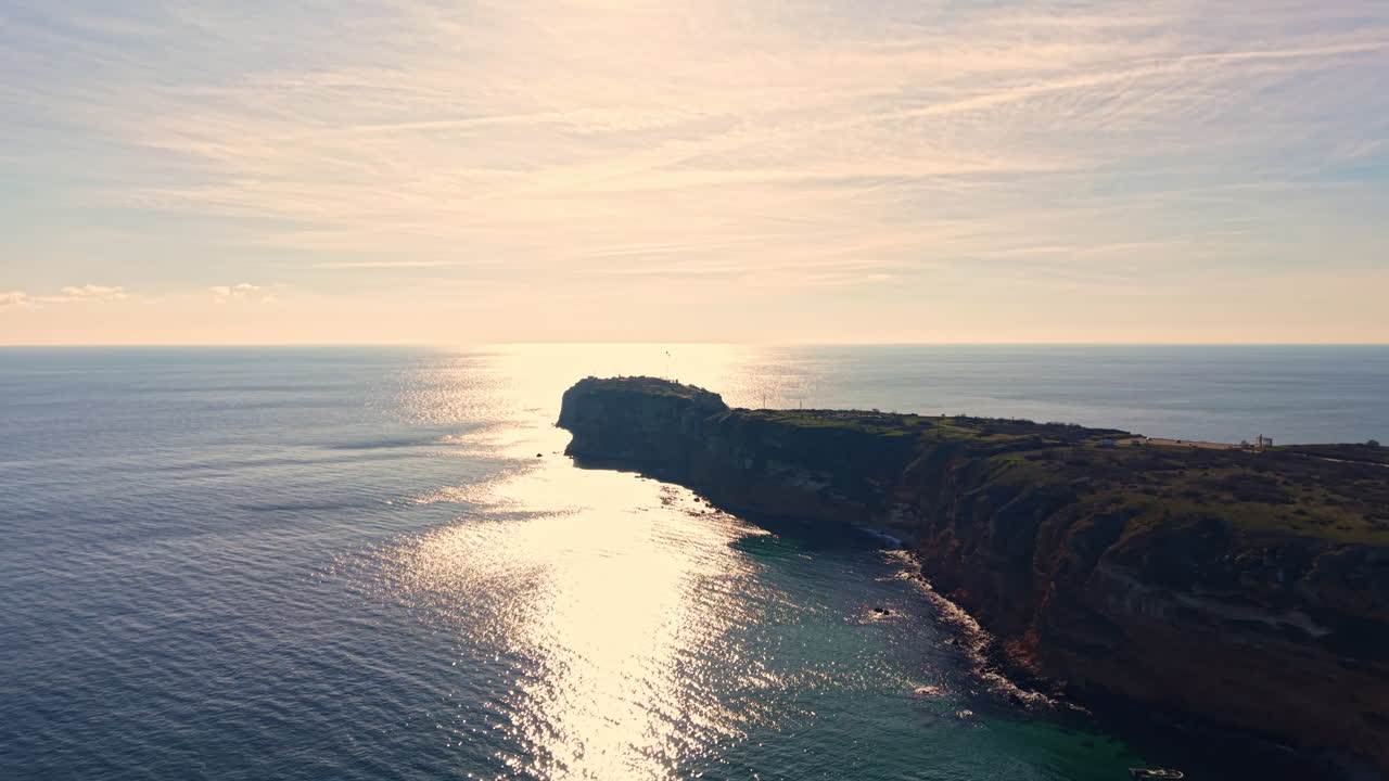 Solnedgang over havet ved en klippekyst med et fyrtårn i det fjerne nær en rolig vandoverflade