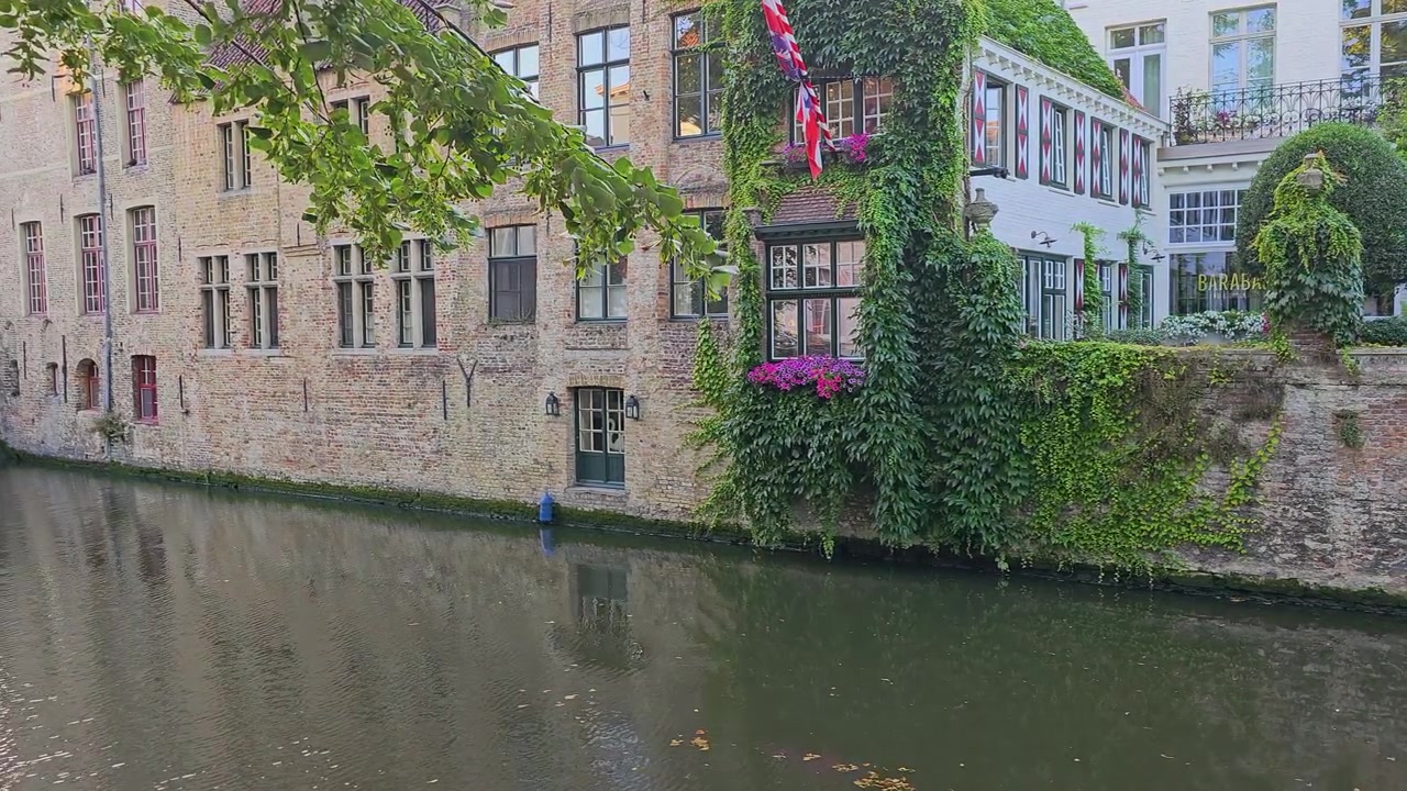 Historisk europæisk kanal med gamle murstensbygninger, buet stenbro, efeu-beklædte mure, løvtræer, blomsterkasser og rolige vandrefleksioner i en charmerende gammel by ved solnedgang.