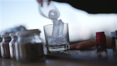 human hand dropping ice cubes into glass with spices and drink preparation tools nearby