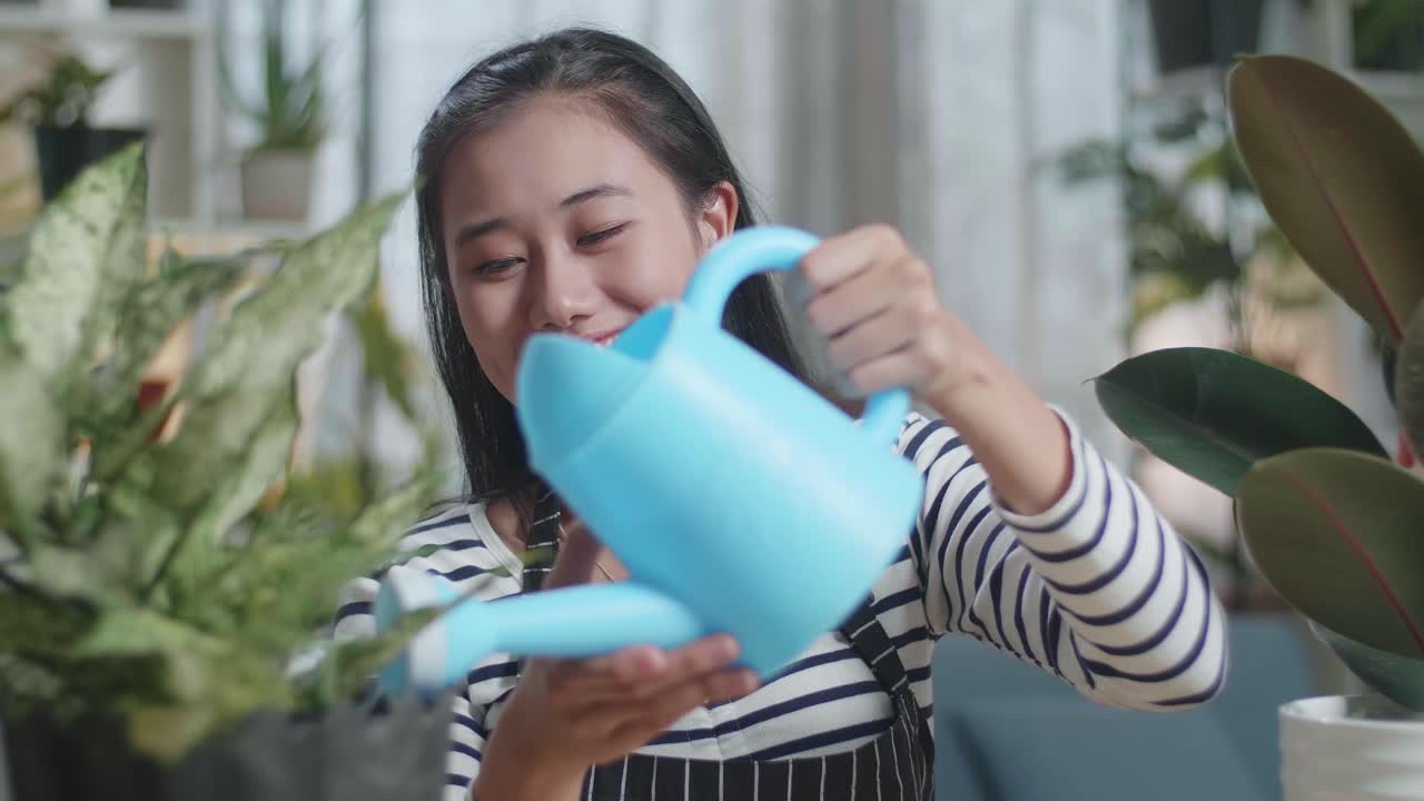 집 에 있는 식물 들 에게 물 을 주기 위해 물병 을 들고 있는 미소 짓는 아시아 여자 의 근접 사진