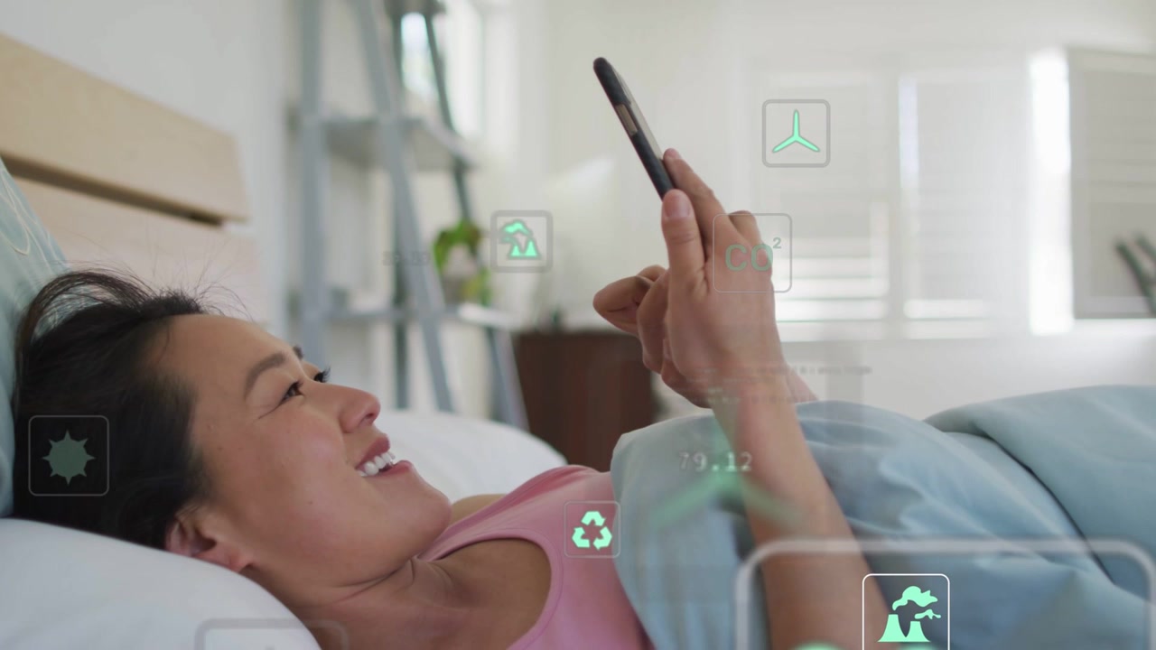 Vídeo vertical: Mulher coreana tocando no smartphone, respondendo a ícones de dados flutuantes na cama, com blusa rosa.