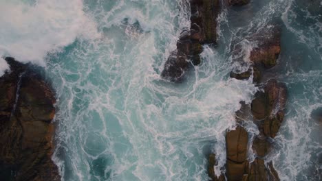 Aerial-view-descending-over-rough-ocean-waves-crashing-and-foaming-against-rocky-coastal-formation