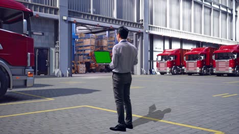 full body back view of an asian male professional worker standing, outside of logistics distributions warehouse, typing on laptop's keyboard with green screen and look at the factory once