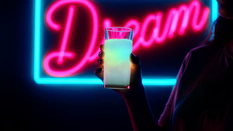 person holding a glowing drink in front of a neon 'dream' sign