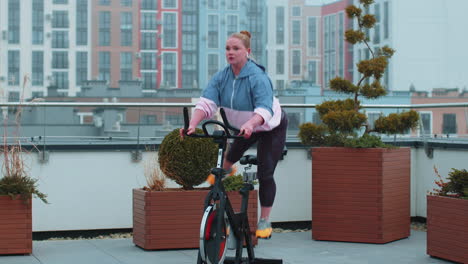 healthy caucasian woman exercising workout on stationary cycling machine bike on house rooftop gym