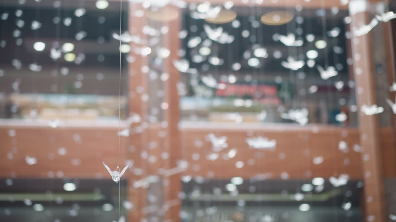 현대 쇼핑몰의 반사 유리 표면의 흐릿한 시야, 초점에 새 모양의 서스펜딩 장식 물체가 특징이며, 배경에는 빛의 동적 보케 효과가 있습니다.