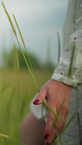 un primer plano de la mano de una mujer con esmalte de uñas rojo, cepillando con gracia contra las hierbas altas en un campo tranquilo y verde. lleva un delicado vestido blanco con patrones, que exuda una atmósfera serena
