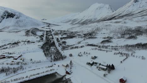Drohnenansicht-In-Der-Gegend-Von-Tromsø-Im-Winter,-Die-über-Eine-Verschneite-Landschaft-Mit-Holzhäusern-Und-Weißen-Bergen-In-Norwegen-Fliegt