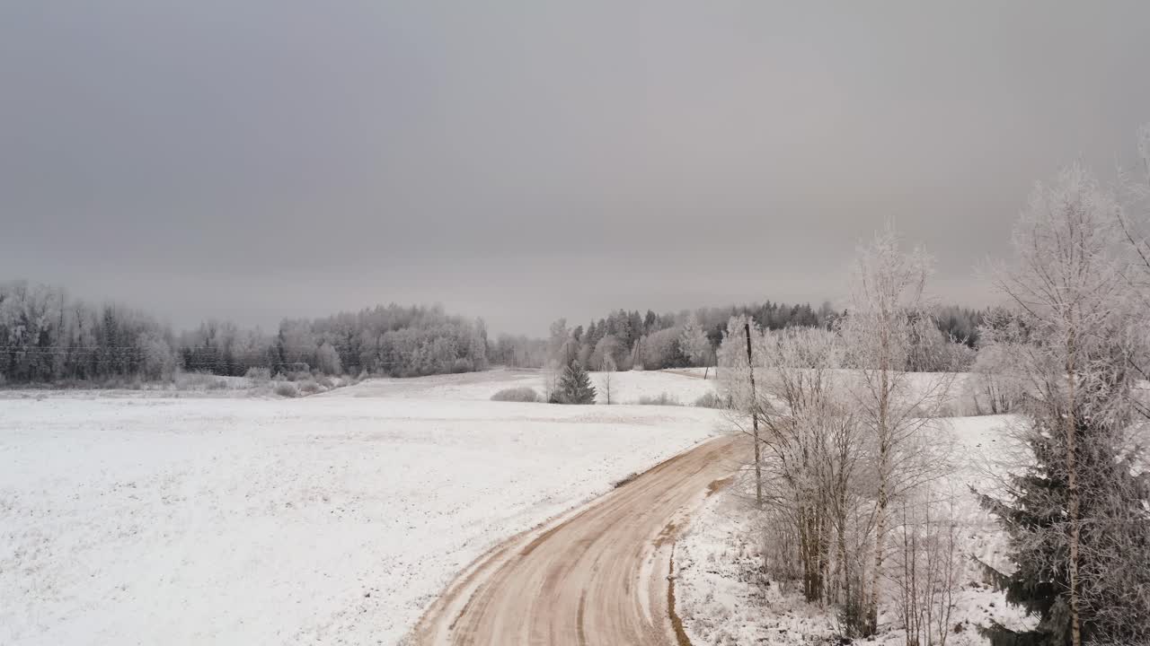 Aerial view of winding gravel road in countryside landscape with snow covered ground and frost in the trees. Winter scenery with frozen forest in the background.