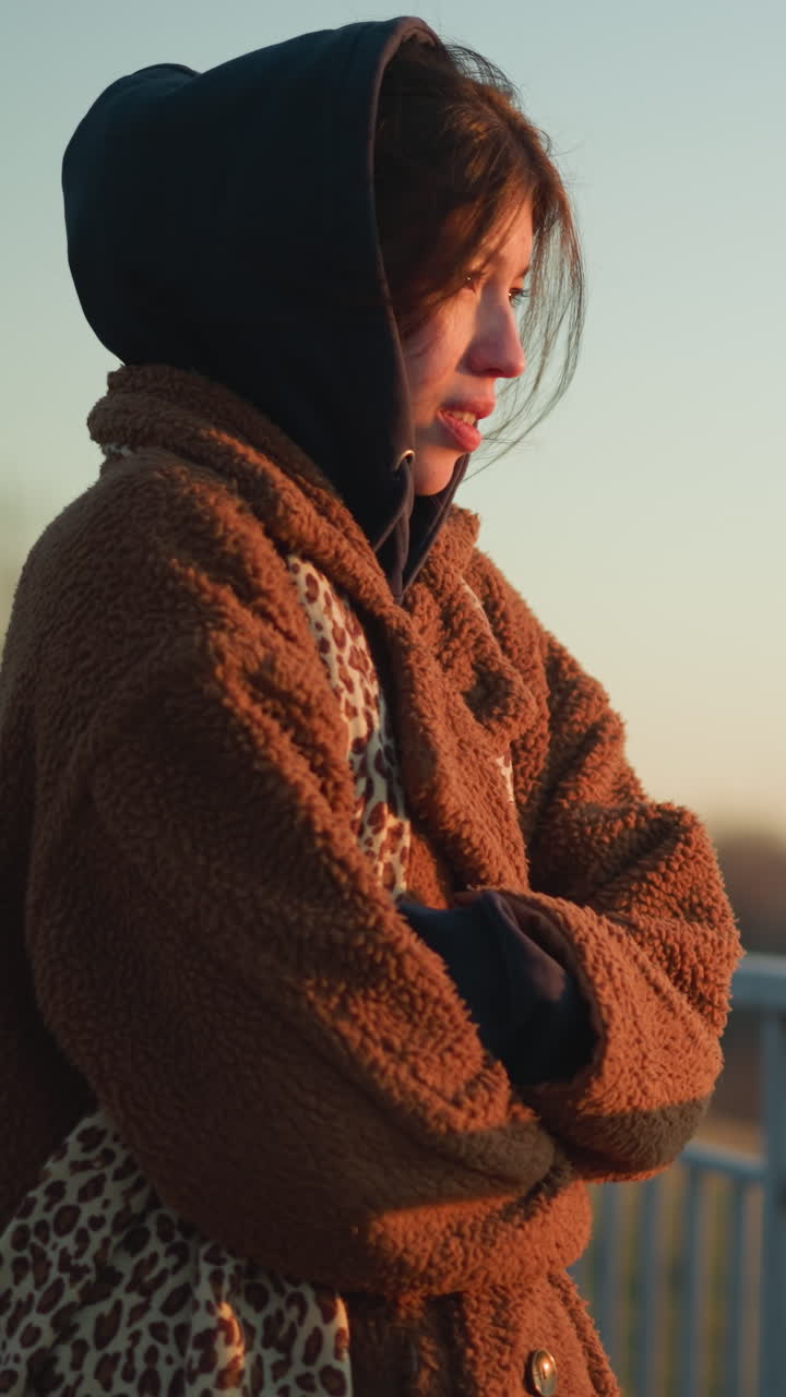 갈색 코트와 후디를 입은 눈물을 흘리는 소녀의 클로즈업, 금속 레일링 근처에 서 있습니다. 그녀의 손은 그녀의 팔에 접혀서 멀리서 사려 깊게 바라보고 슬의 느낌을 반영합니다.