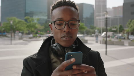 retrato en primer plano de un hombre afroamericano de moda estudiante pasante enviando mensajes de texto navegando usando una aplicación de redes sociales de teléfono inteligente esperando en la ciudad