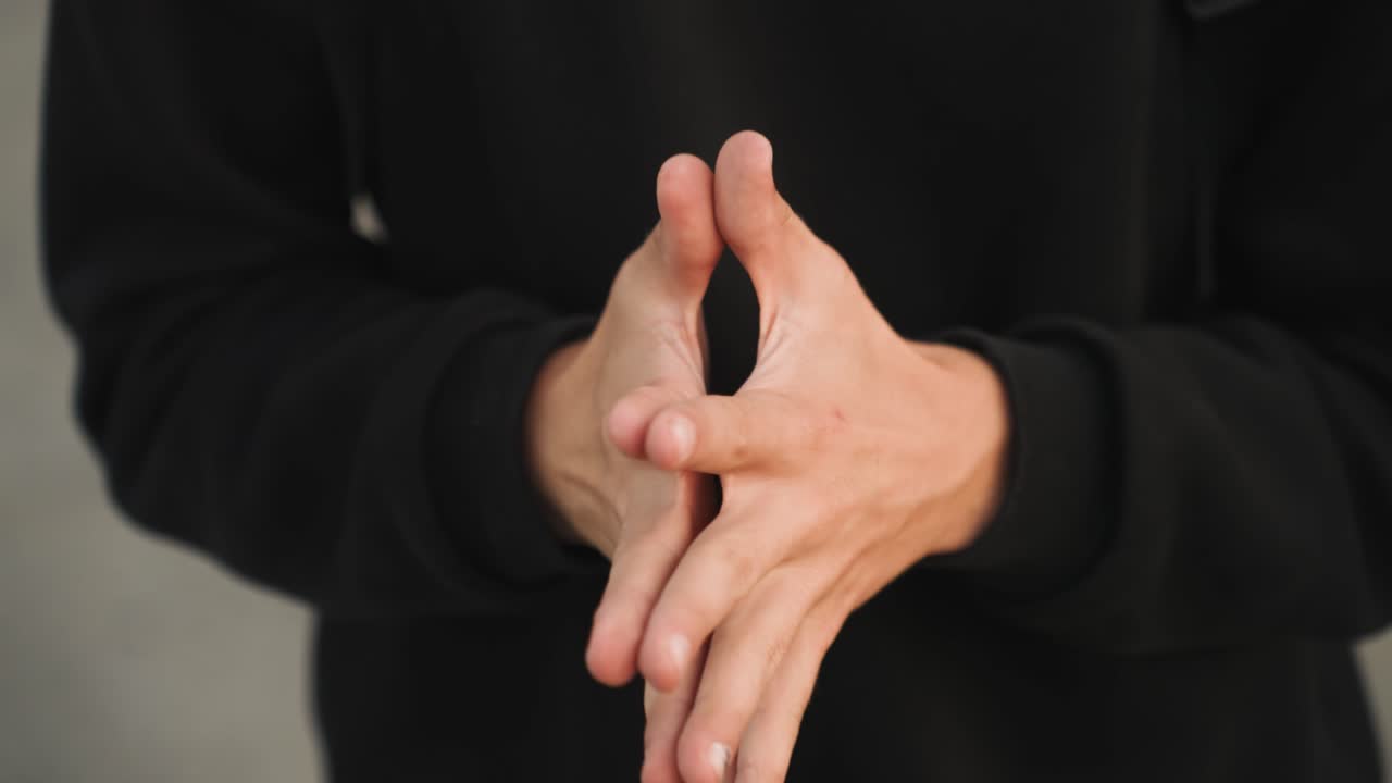 Primo piano di mani che si agitano in una felpa con cappuccio, un giovane adulto si sfrega dita e palmi in un ambiente interno neutro. Dettaglio discreto delle unghie, felpa nera casual. La sequenza mostra gesti calmanti e ansiosi, ideale.