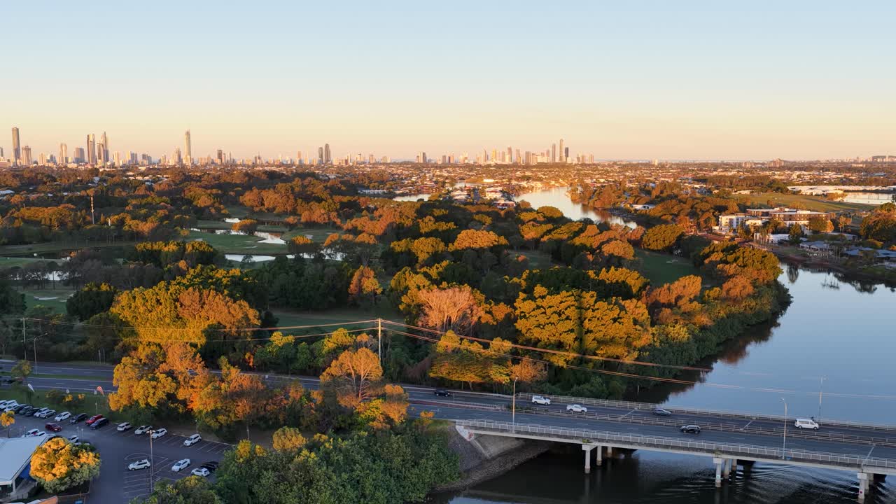 Drone footage glides above a winding river, tree-lined banks, and a busy highway with cars, revealing a distant city skyline under warm sunset lighting