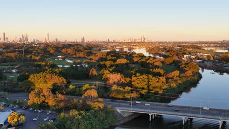 drone footage glides above a winding river, tree-lined banks, and a busy highway with cars, revealing a distant city skyline under warm sunset lighting