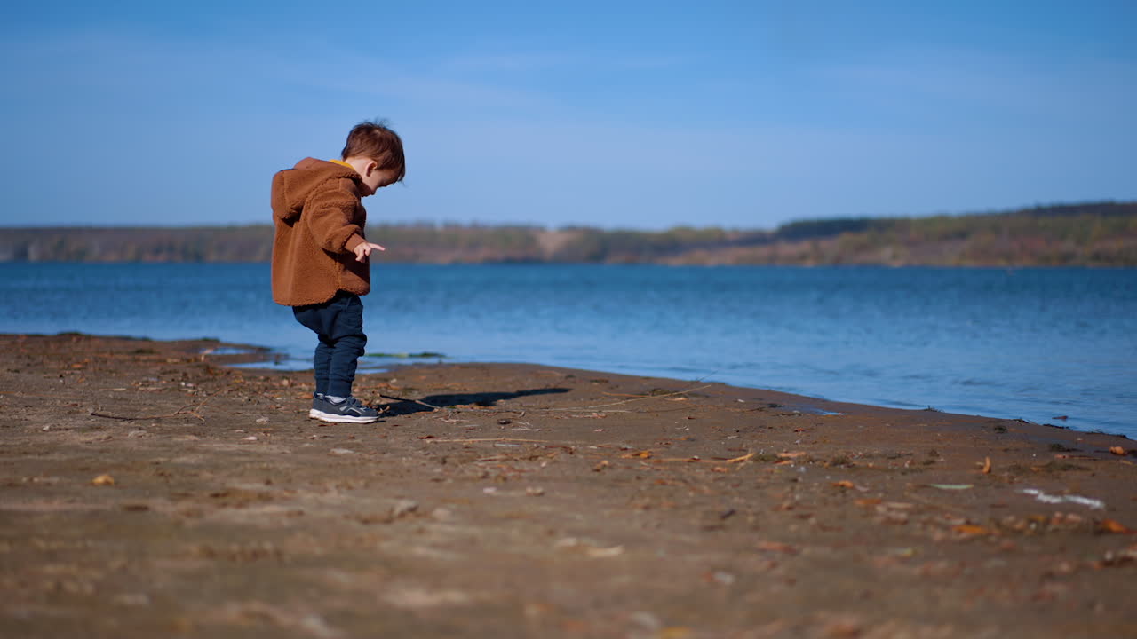 Lovely toddler playing on the river bank on sunny autumn day. Cute baby boy throwing stones into water.