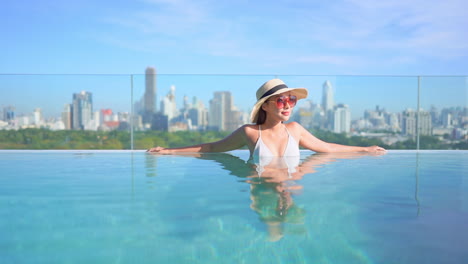 Mujer-Asiática-Rica-En-Piscina-Infinita-En-Un-Día-Soleado-Con-Paisaje-Urbano-En-Segundo-Plano,-Cuadro-Completo