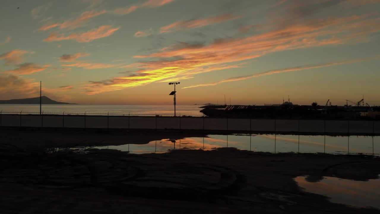Drone nousee ja panoroi Ensenadan rannikkosataman yli, jossa on nostureita ja auringonlaskun heijastuksia.