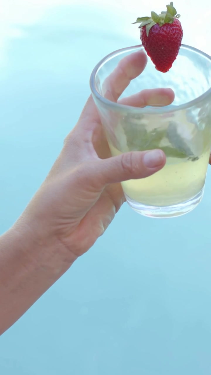 Video vertical: Mujer en traje de baño levantando un vaso con limón y fresa, revelando el cielo sobre la piscina.