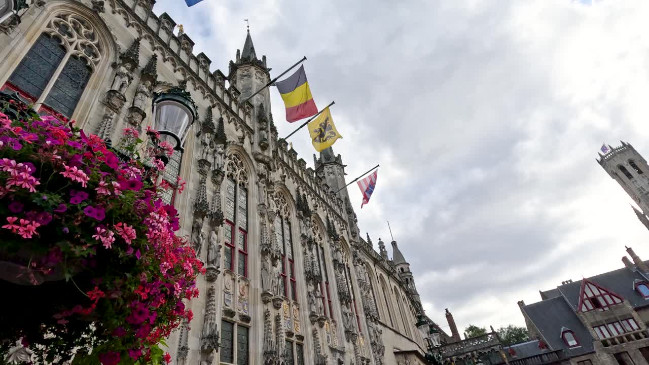 Flags flutter above Bruges City Hall, framed by flowers and medieval architecture on an overcast day