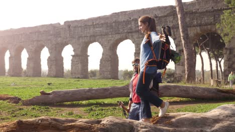 beautiful young woman backpacker walking balancing on a log trunk holding hands with man boyfriend in front of ancient roman aqueduct ruins in parco degli acquedotti park in rome at sunrise romantic sleeping bag slow motion