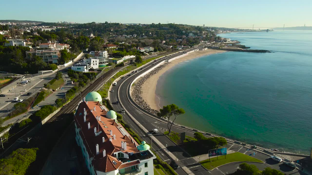 Aerial drone video flying above luxury hotel and apartment Caxias neighbourhood coastal shoreline seaside beach during summer sunny day in Portugal, train and traffic visible near sandy ocean beaches