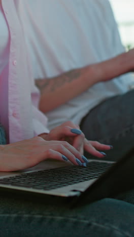 primer plano de una mujer con una camiseta rosa y vaqueros, uñas pulidas escribiendo en una computadora portátil, mostrando algo en su tableta a su colega enfocada a su lado que también está escribiendo rápidamente