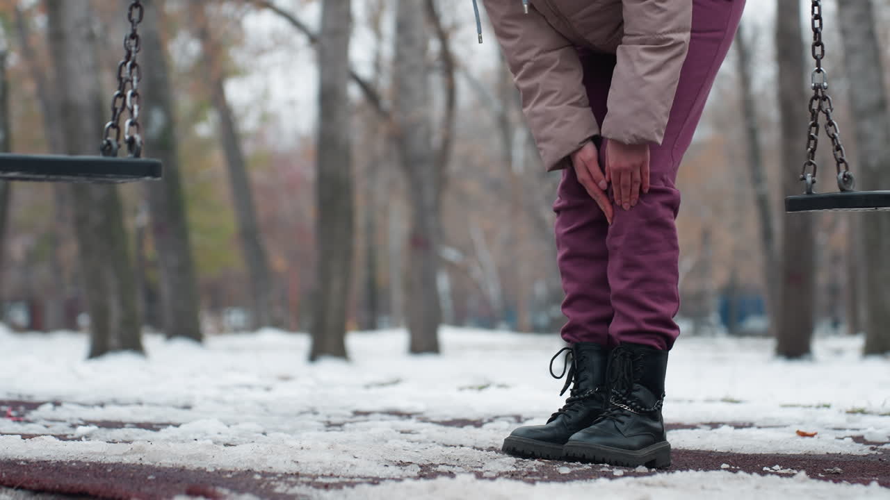 vista de ángulo bajo de una persona joven en traje de invierno sintiendo dolor en la rodilla mientras se dobla y frota la rodilla con un columpio cerca, rodeada de tierra cubierta de nieve y árboles desnudos en el fondo