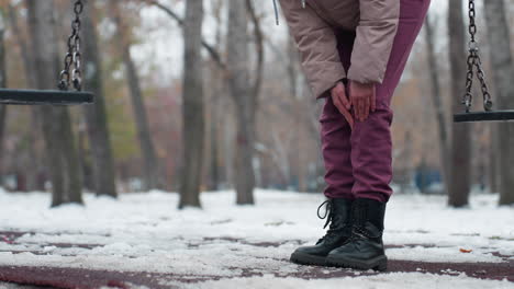 low angle view of young person in winter outfit feeling pain at knee as she bends and rubs knee with swing nearby, surrounded by snow-covered ground and bare trees in the background