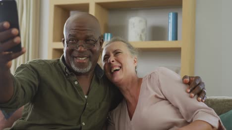 Happy-senior-diverse-couple-wearing-shirts-and-taking-selfie-with-smartphone-in-living-room