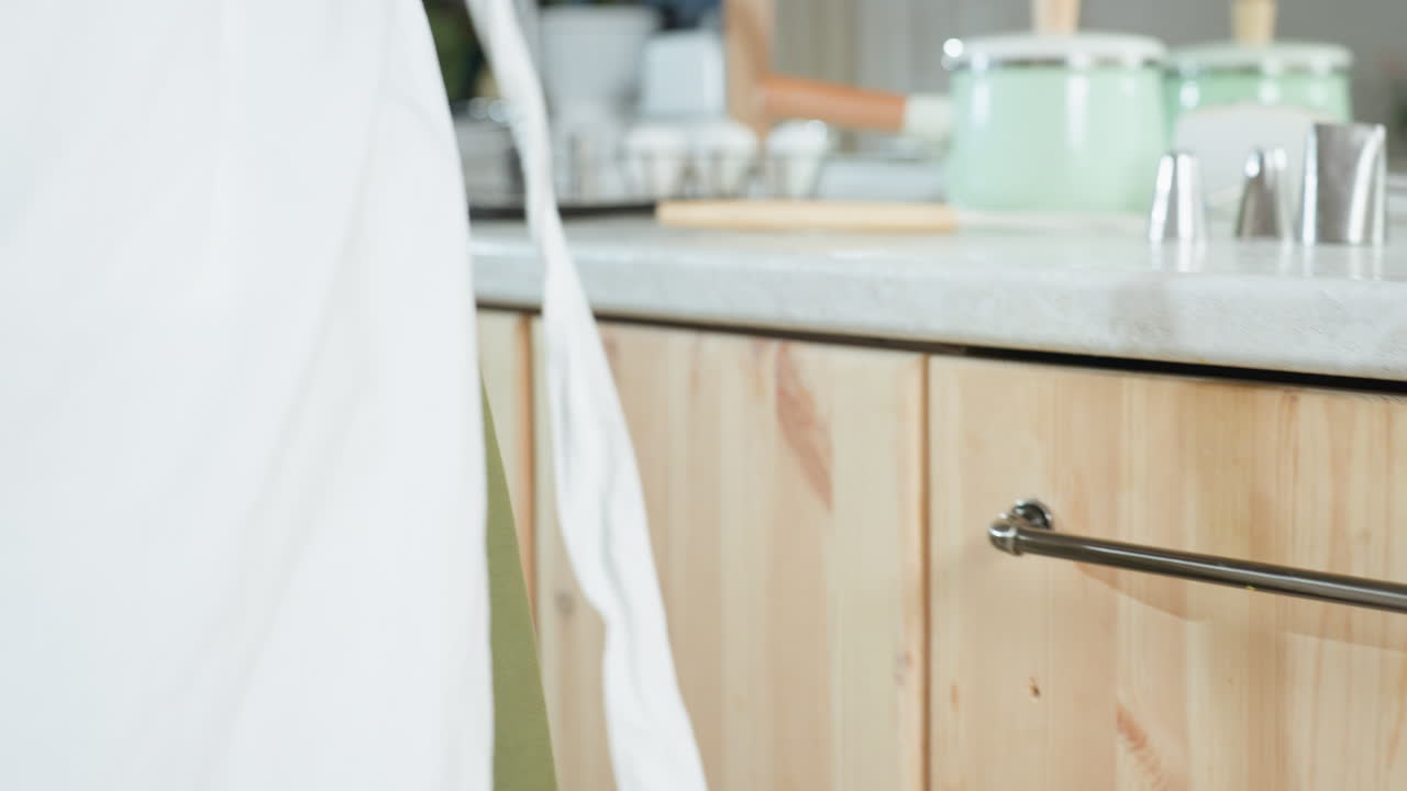 Close up of woman picking up white apron from kitchen drawer handle, preparing to tie it around waist, highlighting gentle hand movement, soft fabric texture, cozy kitchen setup with wooden cabinets