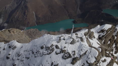 Aerial-top-down-shot-of-summit-cross-on-snowy-peak-with-mountain-lake-in-the-valley-during-sunny-day