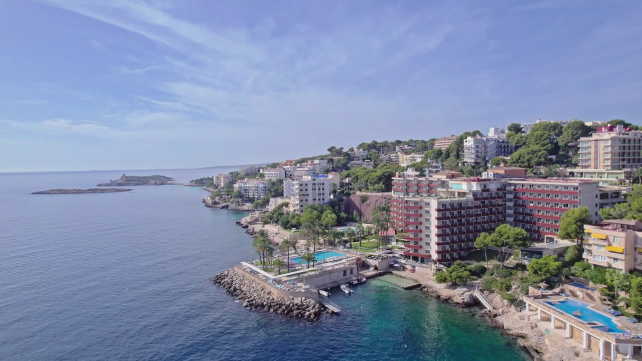 Un drone vola in avanti lungo una costa rocciosa mediterranea, passando accanto a hotel sulla collina, piscine fiancheggiate da palme e un piccolo isolotto roccioso. Acqua turchese calma si estende sotto un cielo azzurro velato
