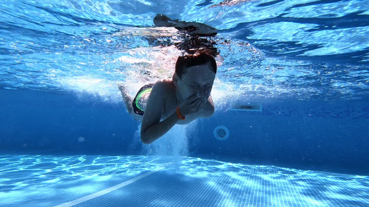 Boy swimming under the clear water in the swimming pool. Young guy jumps underwater and swims with closed eyes and nose on a blue background. Underwater video.