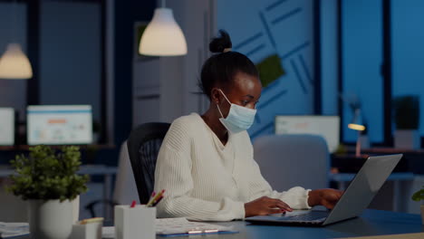 empleado africano con máscara de protección facial trabajando en la computadora portátil horas extras