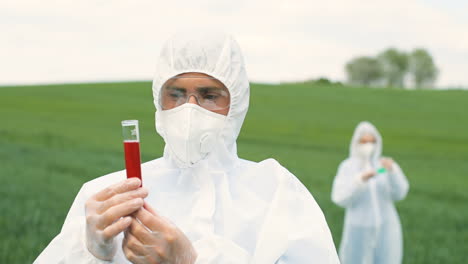 vista de cerca del investigador caucásico con traje protector que sostiene el tubo de ensayo mientras realiza el control de plagas en el campo verde