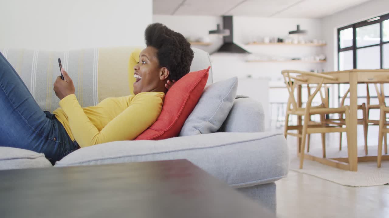 Happy african american woman laying on sofa using smartphone