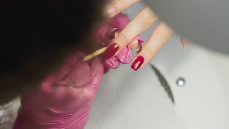 close up view of nail technician wearing pink glove using stick tool to remove excess red nail polish from client fingernail during detailed manicure session under salon lighting