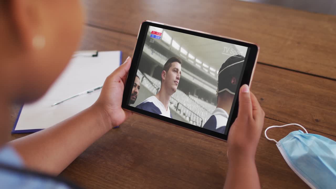 compuesto de una mujer sentada en una mesa con una máscara facial, viendo un partido de rugby en la tableta