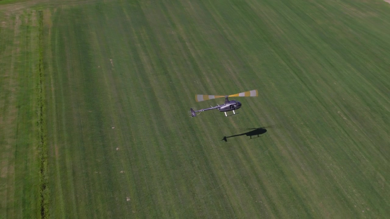 Luftfoto ovenfra af en lille grå helikopter, der flyver fra venstre mod højre og lander på en grøn mark. Dronen stiger op og følger helikopteren, idet den fanger dens tydelige skygge på græsset.