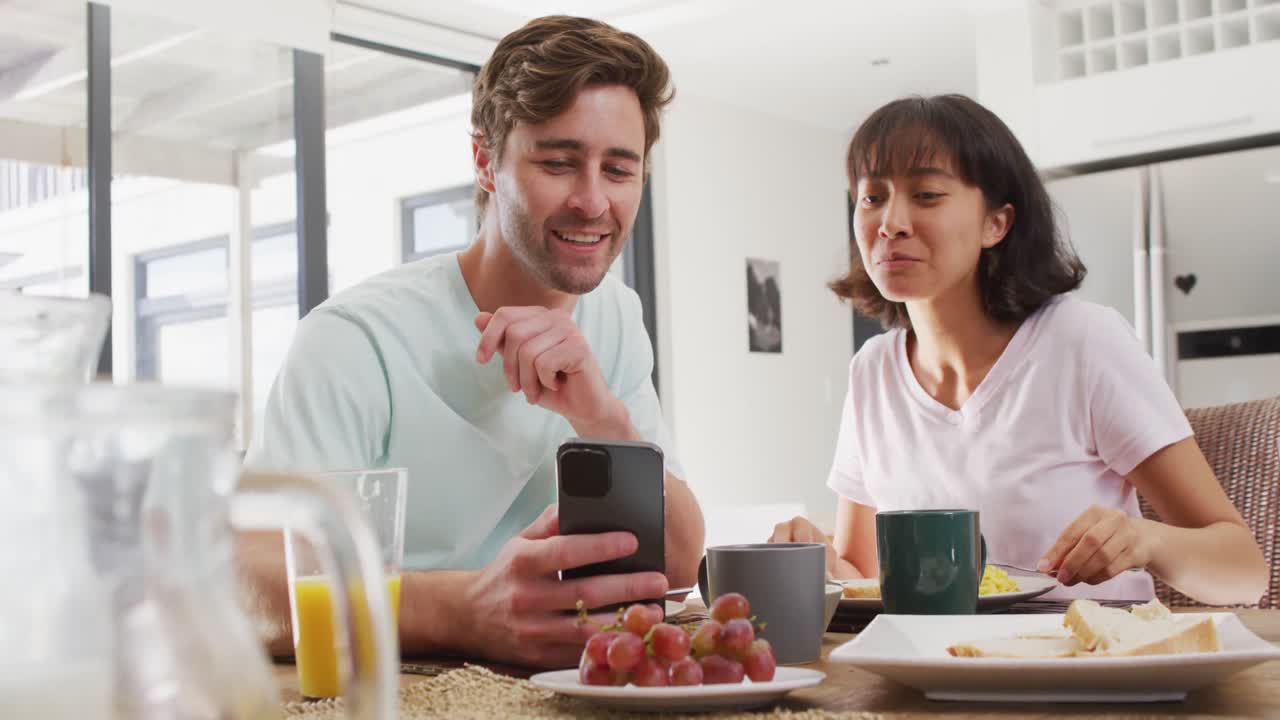 video de una feliz pareja diversa usando un teléfono inteligente y bebiendo café juntos en la cocina