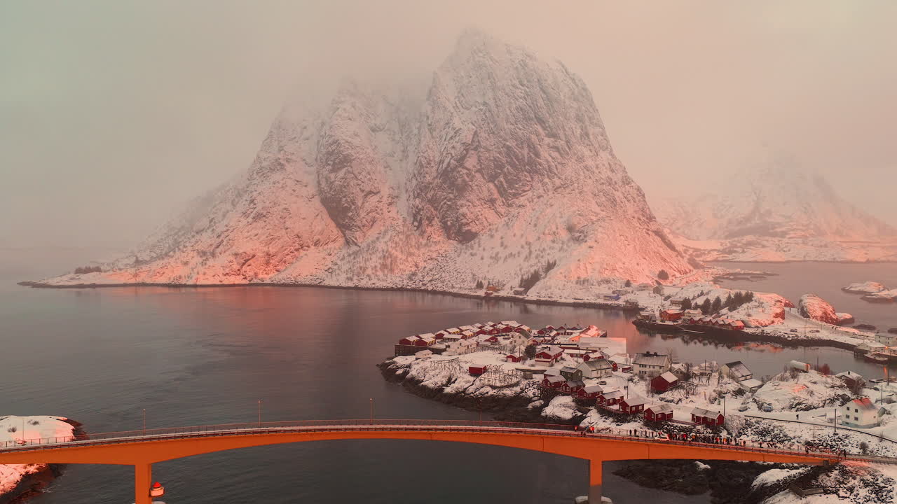 Imponerende luftbilde av Lofoten-arkipelet i Norge, med en bro, tradisjonelle røde hus og snødekte fjell som gløder i solnedgangslyset.