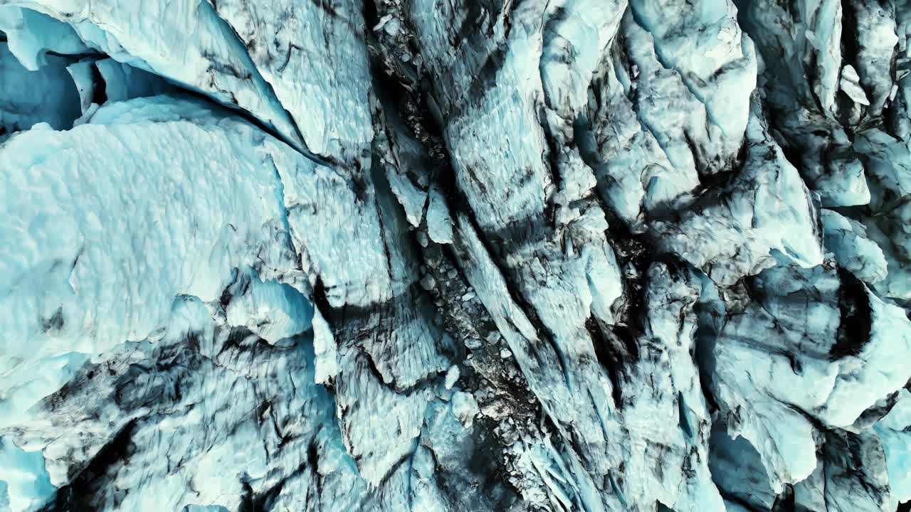 Vista aerea dall'alto di un ghiacciaio blu fratturato con bande di cenere vulcanica scura e stretti crepacci, che creano uno sfondo naturale astratto