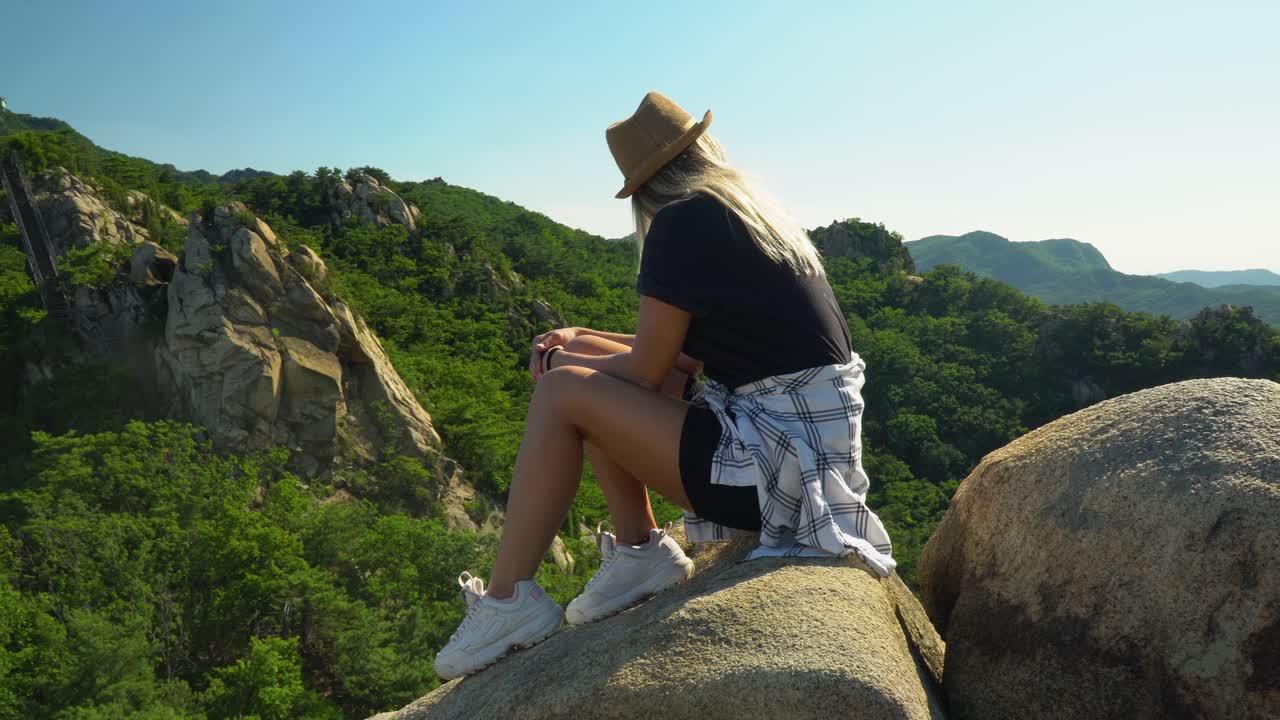chica de moda sentada en las rocas con la mano en la barbilla y admirando el exuberante bosque verde junto a las montañas gwanaksan en un día soleado en seúl, corea del sur