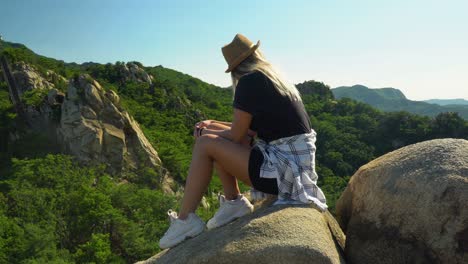 Modisches-Mädchen,-Das-Mit-Der-Hand-Am-Kinn-Auf-Den-Felsen-Sitzt-Und-An-Einem-Sonnigen-Tag-In-Seoul,-Südkorea,-Den-üppigen-Grünen-Wald-An-Den-Gwanaksan-Bergen-Bewundert