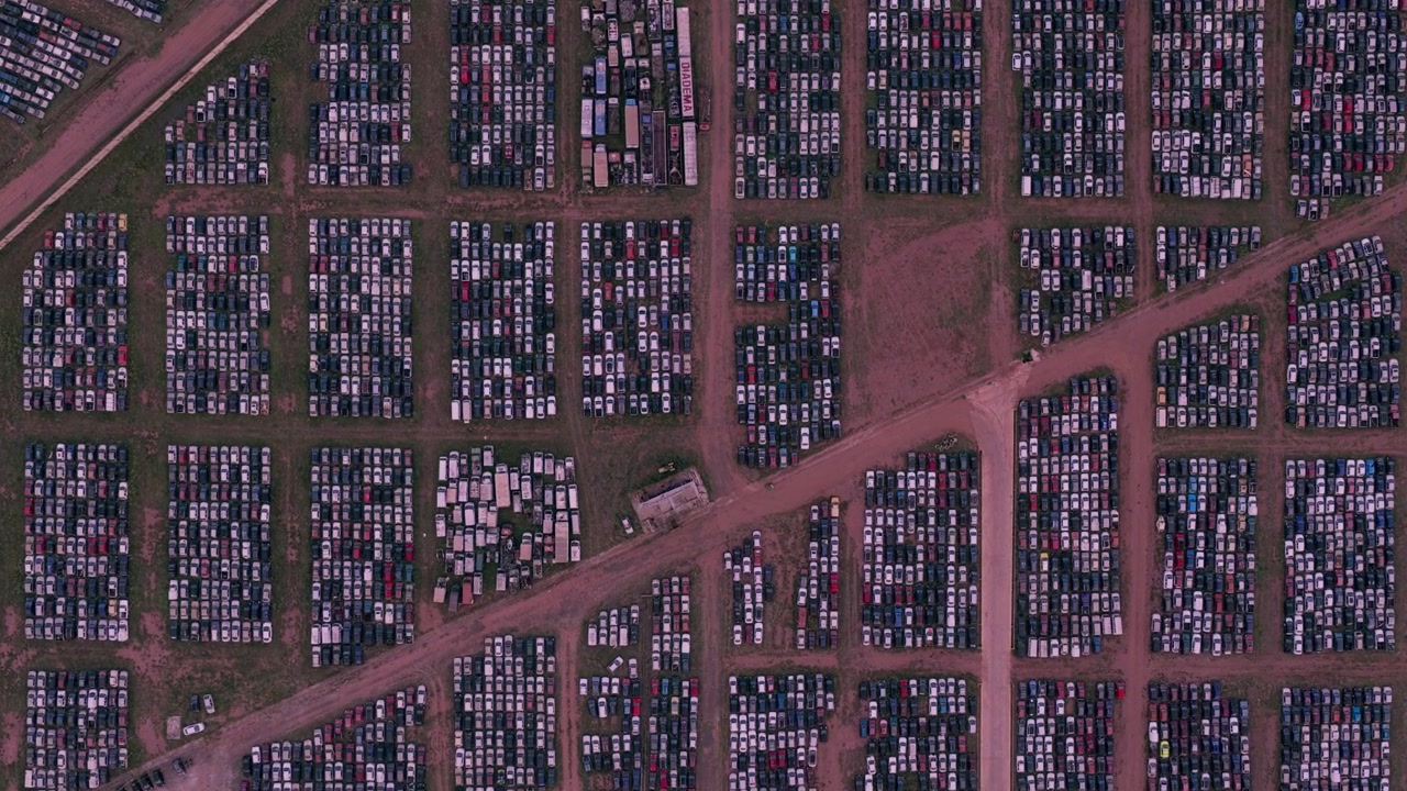 Zoom aereo zenitale su fitte file geometriche di auto parcheggiate all'interno di una vasta struttura di stoccaggio e logistica veicoli