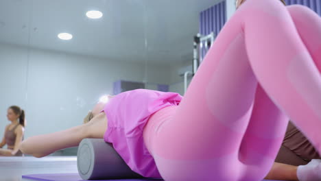 sesión de entrenamiento enfocada con una dama de rosa usando un rodillo de espuma para ejercicios de columna vertebral, con instructor de gimnasia y reflejos en un gran espejo bajo luces brillantes de gimnasio
