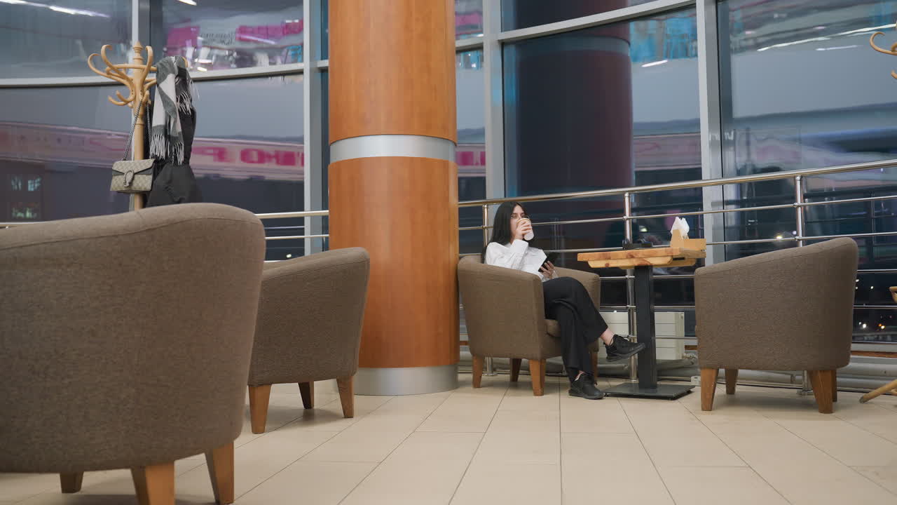 woman sips drink then places it on table while focusing on phone in cozy mall lounge with armchairs, wooden table, tissue box, window reflections and coat rack visible in soft modern background