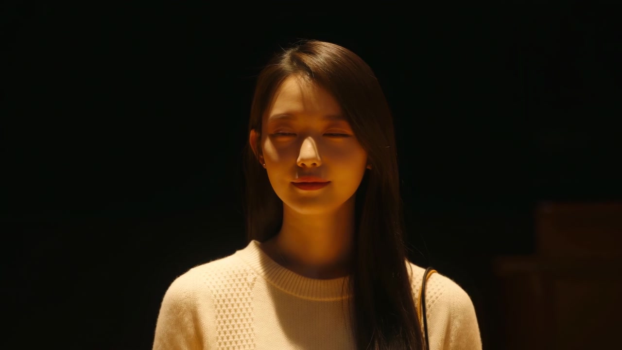 正面を向いた女性が、前を見ていた状態から目を閉じて休む姿勢に変わり、深い影を背景に、彼女の顔と薄手のニットトップが温かい指向性のある光に照らされています。