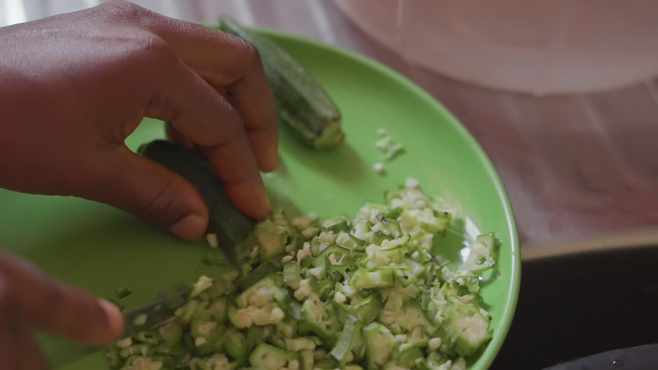 Manos en primer plano picando okra en plato verde, ritmo suave, cocinero casero corta hábilmente okra fresca en trozos pequeños en un plato verde brillante, luz cálida de cocina, enfoque en la textura y el movimiento, íntimo doméstico.